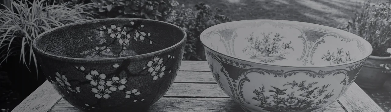Two bowls on a garden table: white cherry blossom on a dark crackle glaze, and French-style floral cartouches with birds on white